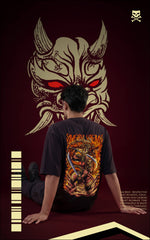 Back view of a model sitting on the floor wearing a black graphic tee with a large, fiery samurai and dragon illustration. A giant stylized Hannya mask with glowing red eyes is superimposed in the background.