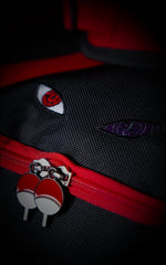 Detailed macro shot of embroidered Sharingan and Rinnegan eyes on black canvas fabric, positioned above dual Uchiha crest zipper pulls on a red track.