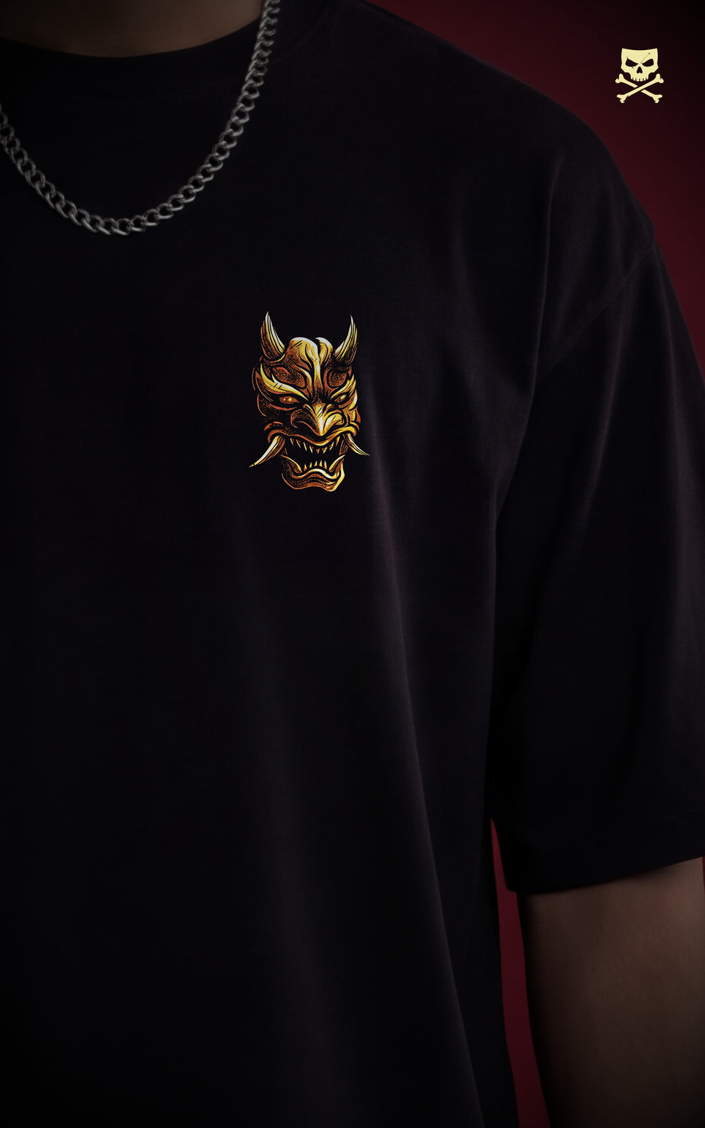 
Close-up of a small, high-detail chest print on a black streetwear shirt depicting a golden Hannya mask. A silver cuban link chain is visible at the neckline, highlighting a minimalist urban fashion aesthetic.