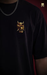 
Close-up of a small, high-detail chest print on a black streetwear shirt depicting a golden Hannya mask. A silver cuban link chain is visible at the neckline, highlighting a minimalist urban fashion aesthetic.