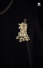 Detailed close-up of a pocket-sized chest print on a black t-shirt. The graphic depicts a toxic waste bottle sitting on a pile of skulls with prominent Japanese kanji characters "中毒" (toxic/poison) overlaid in a cream-colored, distressed style.