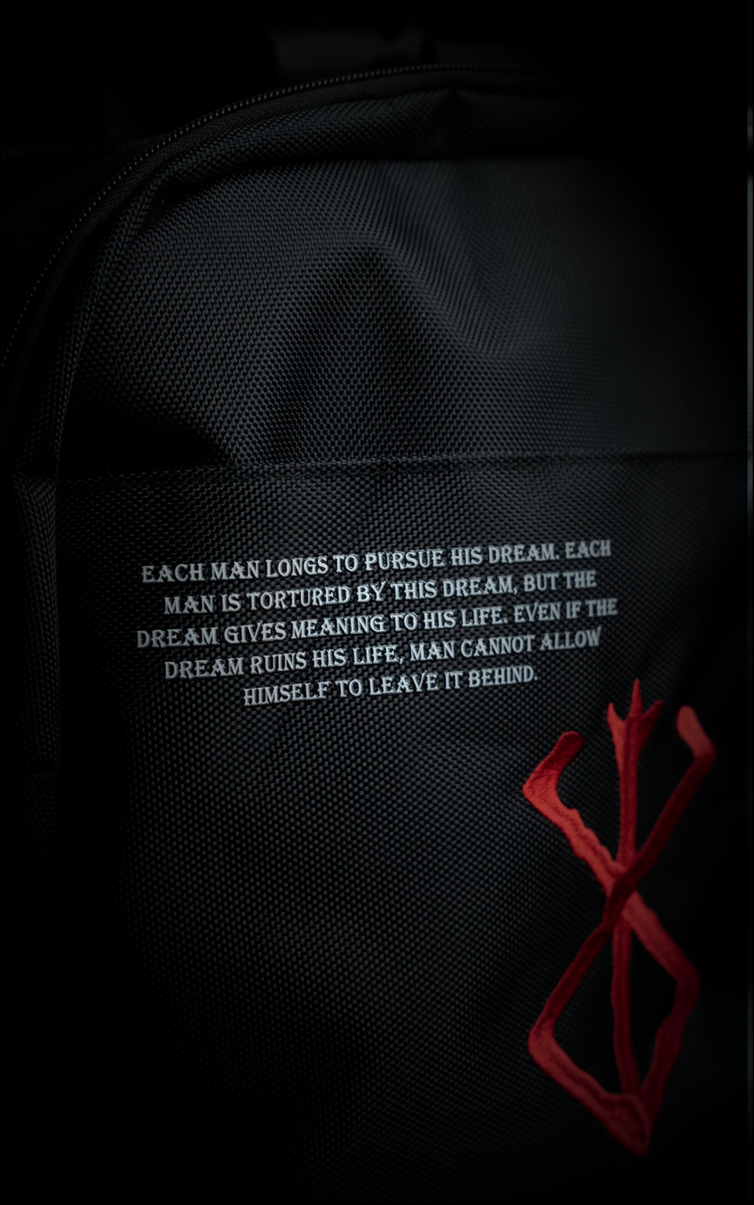 Close-up of the Berserk duffle bag showing Griffith’s "Each man longs to pursue his dream" quote in white text next to an embroidered red Brand of Sacrifice symbol.
