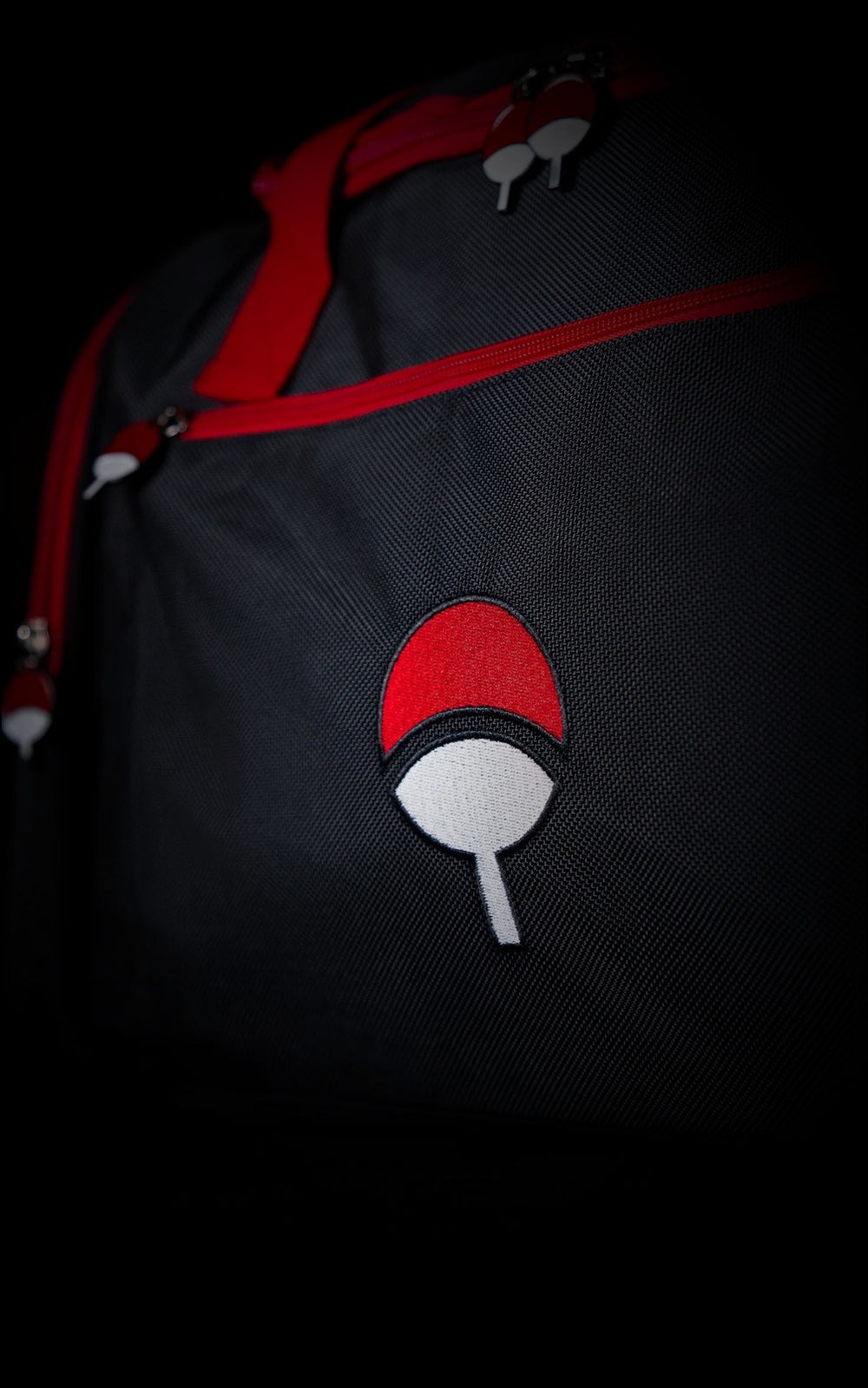 Close-up of a black anime gym bag showing a high-quality embroidered red and white Uchiha clan fan symbol and custom enamel zipper charms.