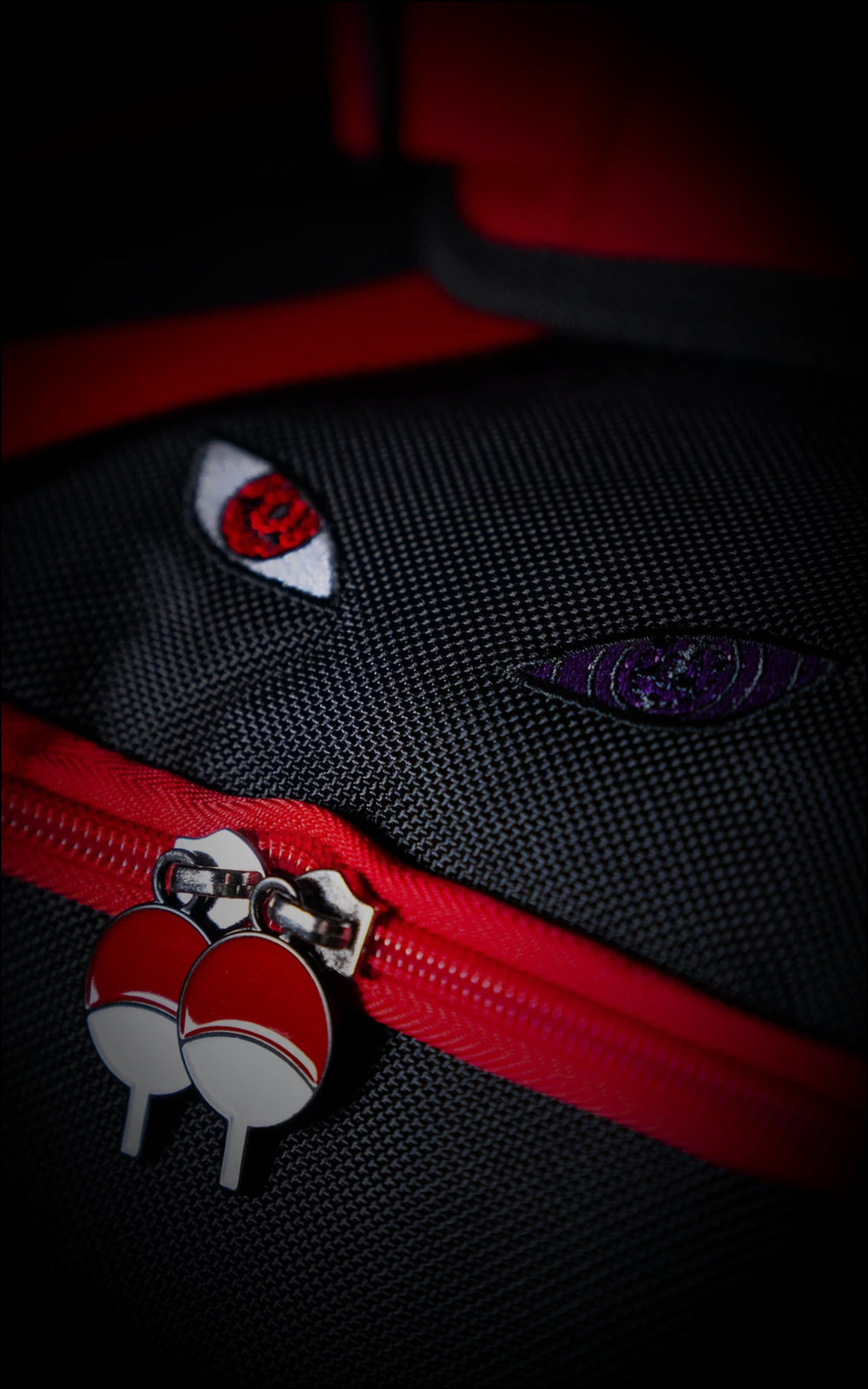 Detailed macro shot of embroidered Sharingan and Rinnegan eyes on black canvas fabric, positioned above dual Uchiha crest zipper pulls on a red track.