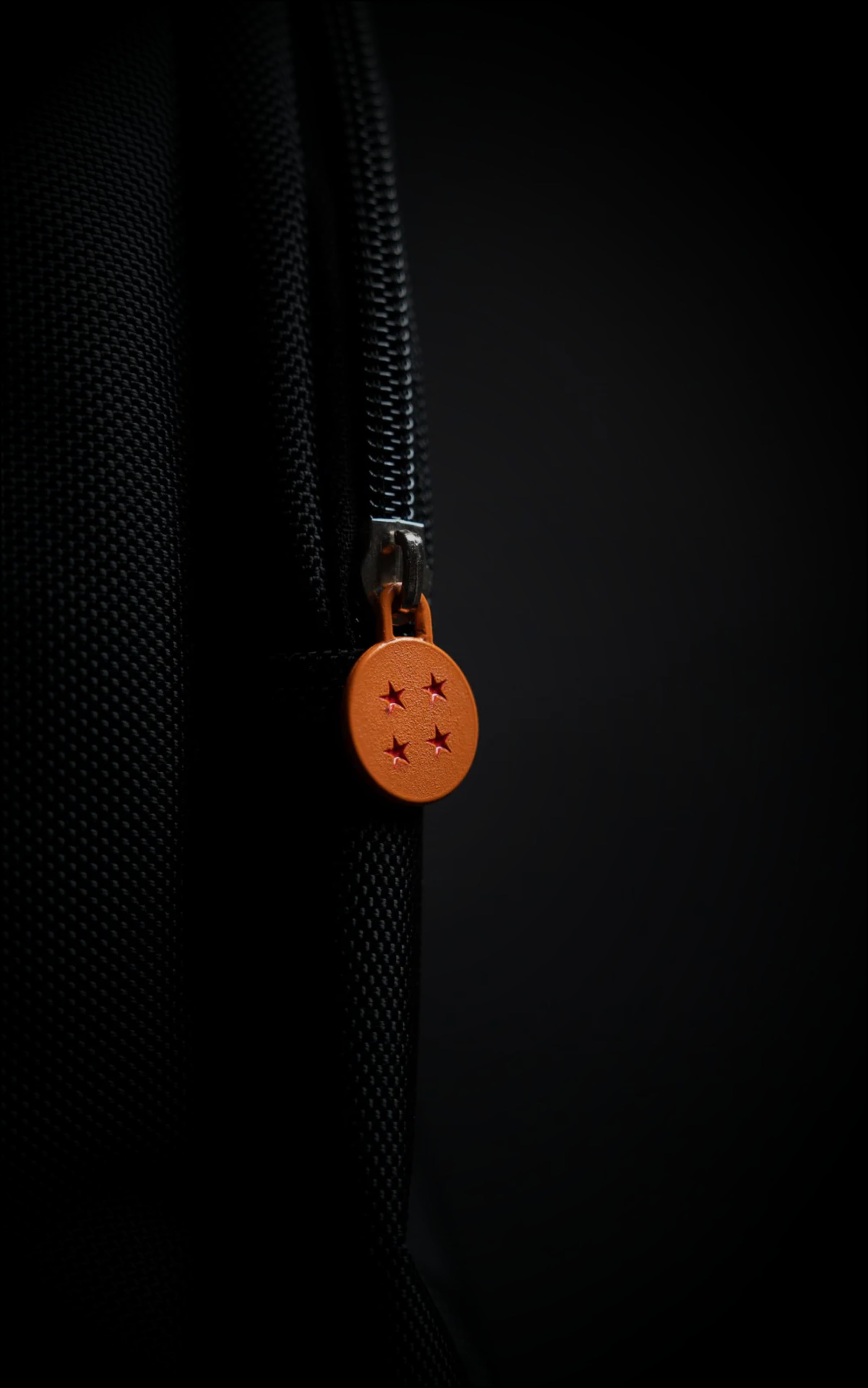 Macro shot of a custom orange circular zipper pull designed to look like the iconic 4-star Dragon Ball with red star engravings on a black zipper track.
